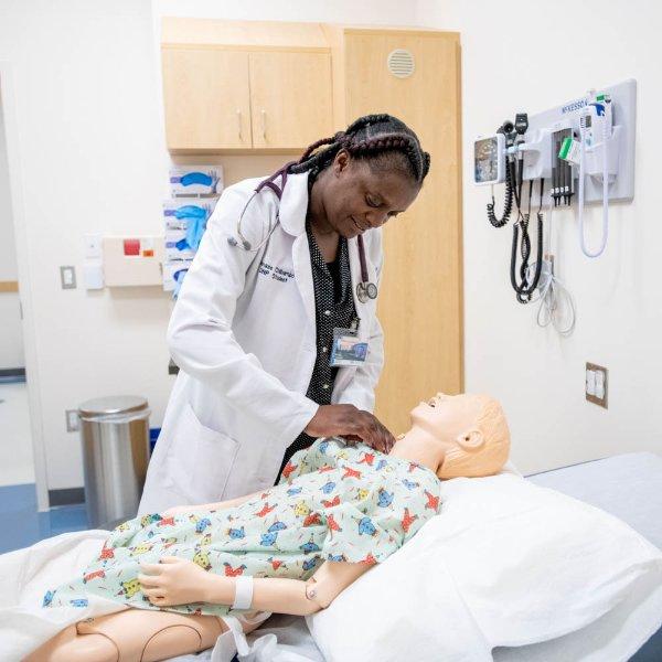 nursing student in white lab coat assesses a manikin that is in a hospital bed