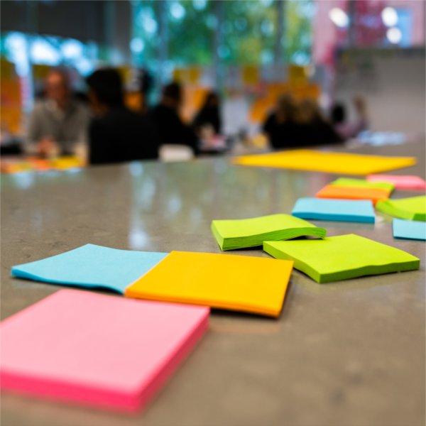 Several pads of colorful sticky notes are scattered on a table. People are in the background.