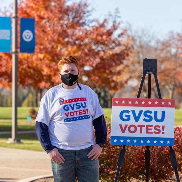 Sam Jacobs in mask standing outside next to GVSU Votes sign and wearing the same t-shirt, with GVSU Votes!