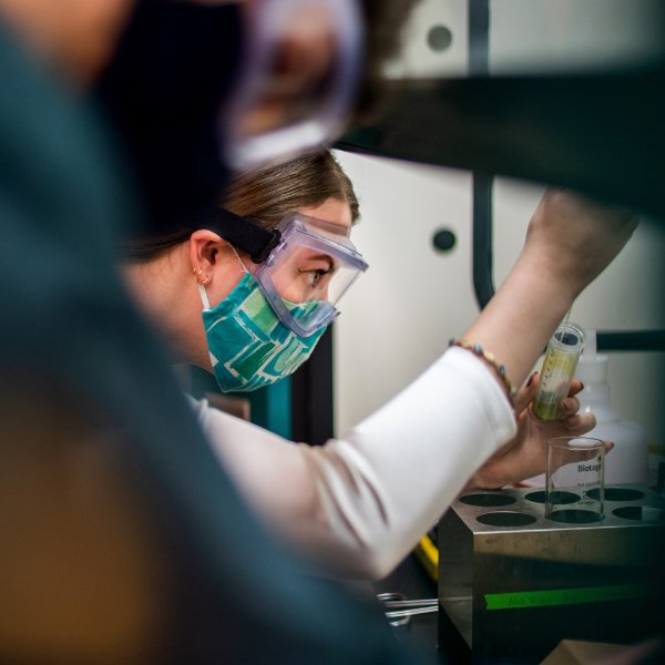 A student wearing goggles and a mask works with a test tube.