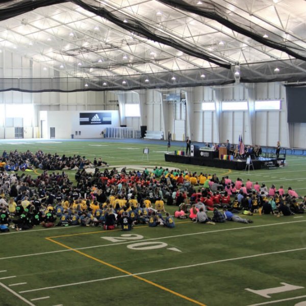 A large group of people sits on a floor with football-like yard markers. There also is a podium where people are sitting and one person is talking.