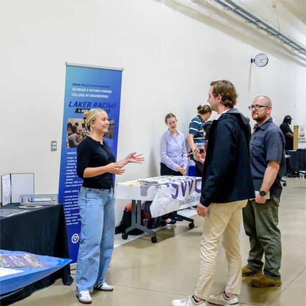two people standing in hallway talking to engineering student, who is standing by the Laker Racing car