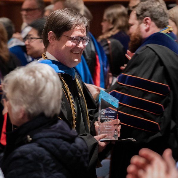 Robert Adams holds an award in a crowd of people, some are in academic regalia