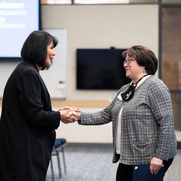President Mantella, left, shakes the hand of Betty Schaner. Both people are standing in a room.