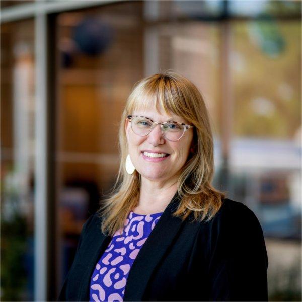 A portrait of Holly Jasinski, a woman with blonde hair and glasses. She smiles and poses in front of the Center for Women and Gender Equity.