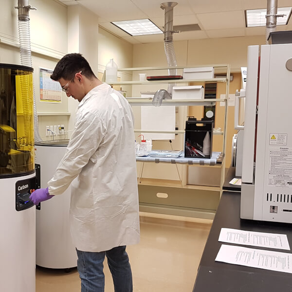 A technician in a lab coat stands in a lab in front of a Carbon, Inc., 3D printer. The printer is a tall cylinder with an amber plastic shield in front of it.