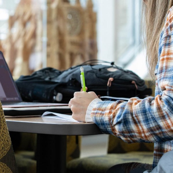 A person's hand holds a pen while the person writes on paper. A backpack and laptop are also on the table.