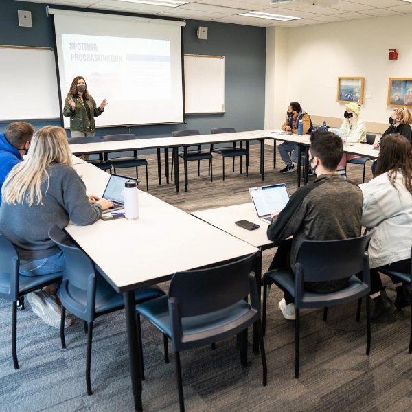 Jessica Fillmore leads a small group workshop, standing and speaking at the front of the room, students in chairs at tables shaped like a box. Screen reads Spotting Procrastination
