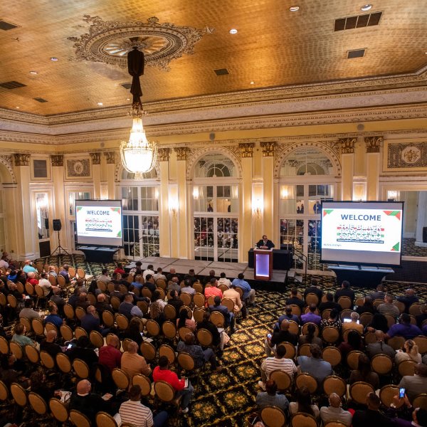 large crowd in ballroom seated with speaker and podium at front, 2 projection screens, large light fixture hanging from ceiling