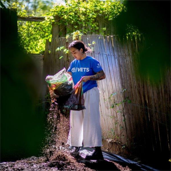 Alex-Mari Ford works on spreading mulch in a yard. She stands next to a fence and holds a bag of mulch.