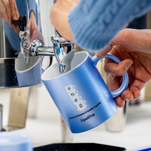 A person pours tea from a coffee urn into a blue mug with text on it that reads �Center for Women and Gender Equity, Equali-Tea�.
