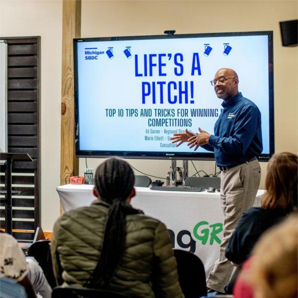 Audience facing a presentation titled "Life's a Pitch! Top 10 Tips for Winning Competitions." A speaker stands by the screen; another by a whiteboard.