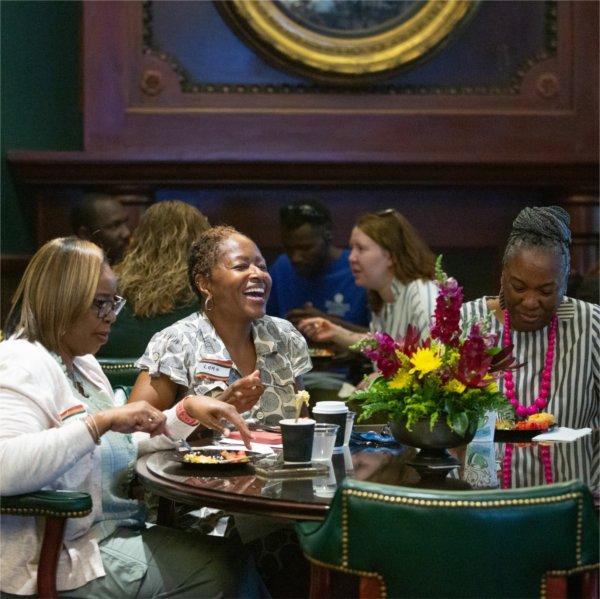 Three well-dressed women with name tags on sit at a table and laugh together as they eat breakfast.