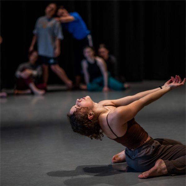 A dancer is on their knees, bending back with arms reaching out. Other dancers are in the background.