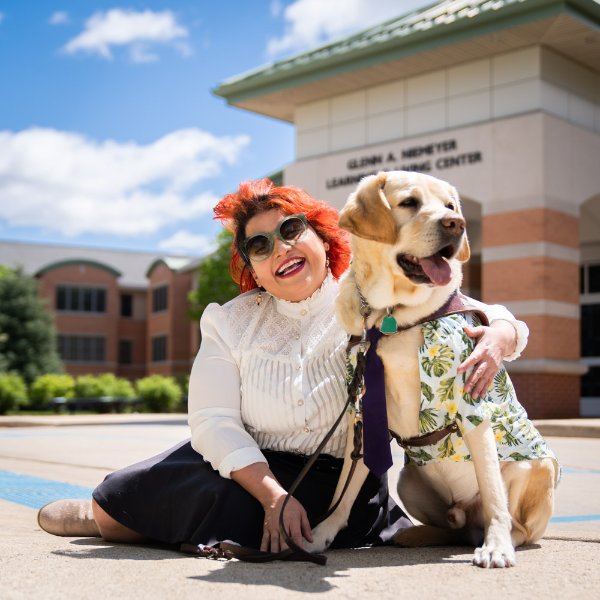 Melba Vélez Ortiz sitting on the ground in front of Frederik Meijer Honors College with her guide dog, Chad.