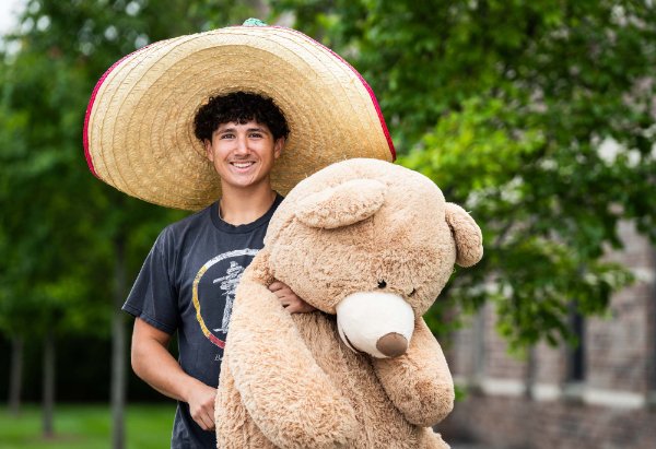 A college student wearing an oversized hat holds a large stuffed teddy bear.  