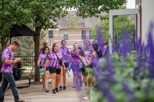  A group of college students wearing matching purple tie-dyed T-shirts walk on a college campus. 