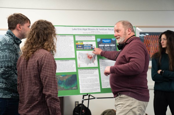 A person points to a poster containing information on research about Lake Erie algal blooms while talking with others.