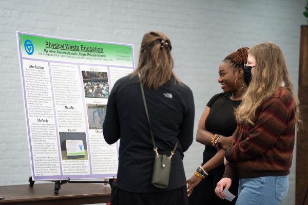 Three people, one of them smiling, one wearing a mask, one with back to the camera, looks at a research board.