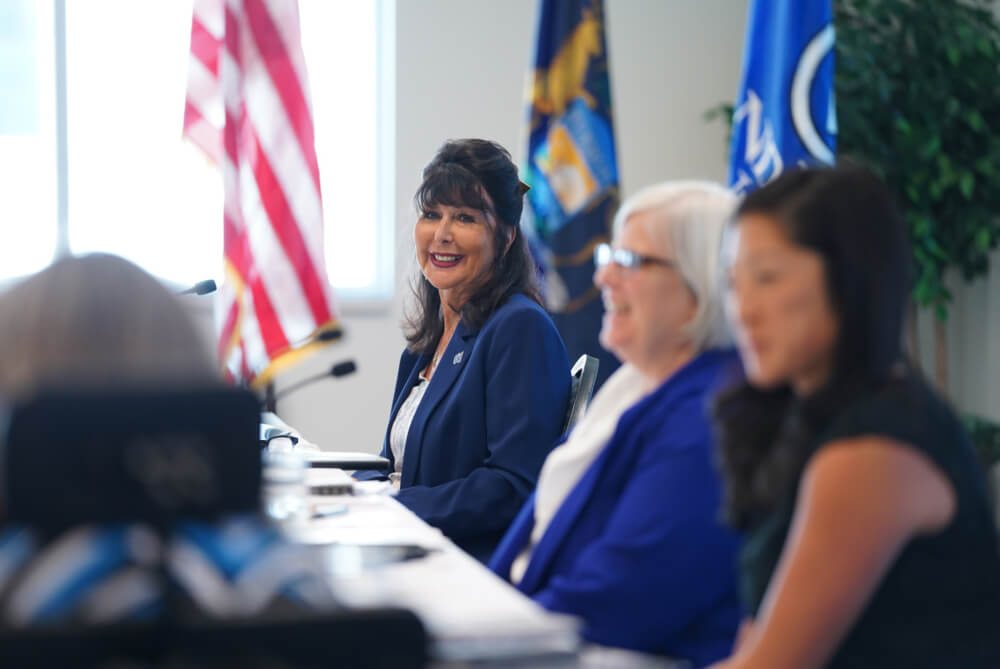 President Mantella sits at a table with the GVSU flag behind her, with Board Chair Mary Kramer and Vice Chair Megan Sall to her left.