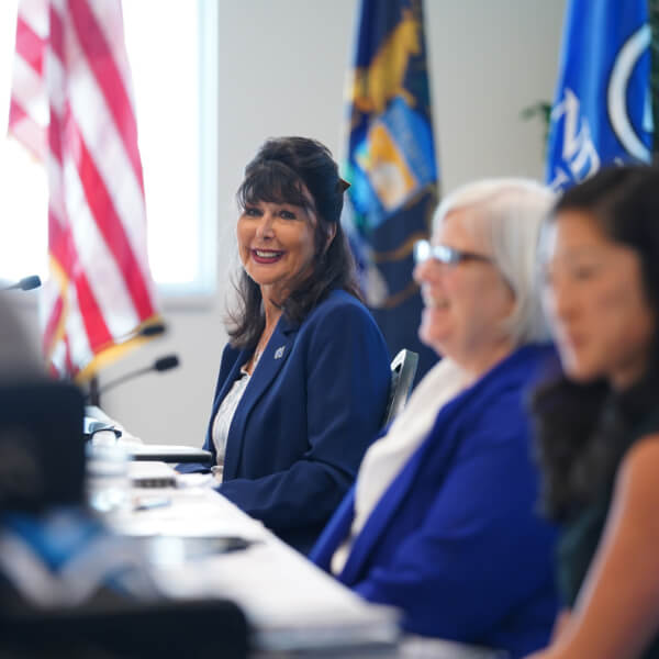 President Mantella sits at a table with the GVSU flag behind her, with Board Chair Mary Kramer and Vice Chair Megan Sall to her left.