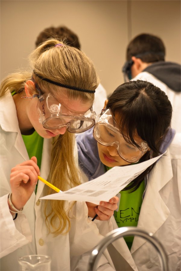Two people wearing goggles peer at a piece of paper held by one person. That person also is holding a pencil.