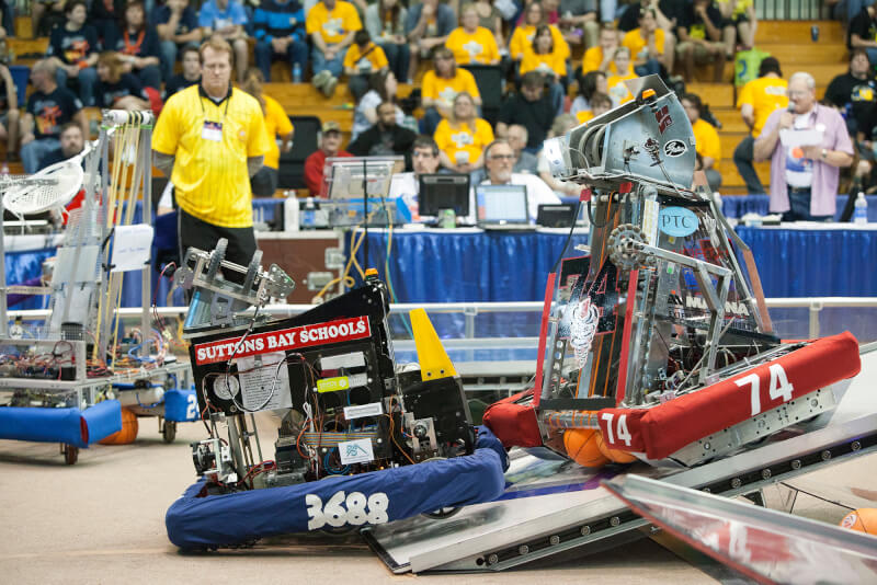 Two robots work to complete an obstacle at a previous FIRST robotics tournament in 2012. They are climbing over a small wall.