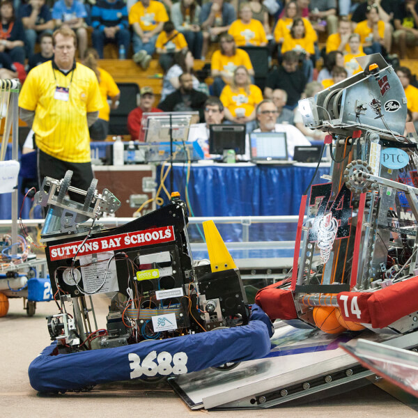 Two robots work to complete an obstacle at a previous FIRST robotics tournament in 2012. They are climbing over a small wall.