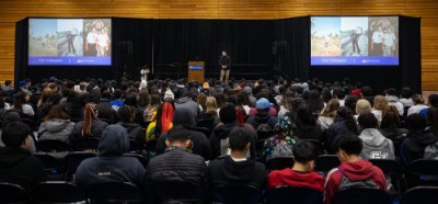 Fieldhouse full of students seated on chairs, watching presentation by man on stage, with two projections on either side of stage
