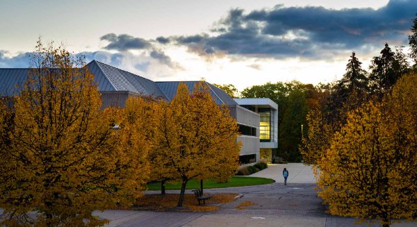  A student wearing a blue backpack walks through campus surrounded by colorful fall leaves and a cloudy sky