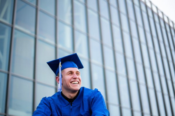 A person wearing a blue cap and gown smiles while looking off to the side. The side of a building with many windows is in the background.