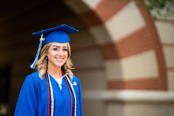A person wearing a cap and gown smiles. A brick arch is in the background.