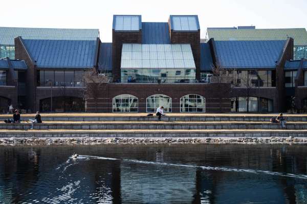 A duck on a pond with a person sitting on a tiered area is the foreground to a building.
