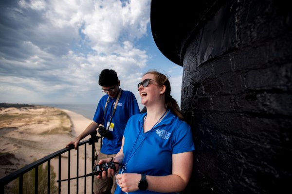  Two people stand by a railing at the top of a lighthouse. One is holding a portable radio and is laughing, while the other is looking down. 