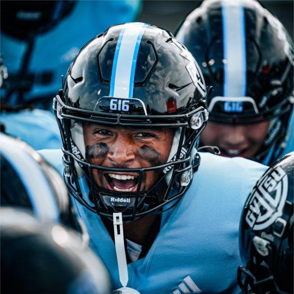 A Grand Valley football player wearing a helmet and light blue jersey fires up his teammates as they crowd around him.ammates.