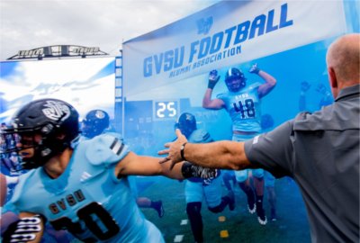 Grand Valley football players run under a banner and through a cloud of blue smoke and the start of a game. 