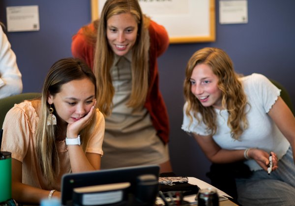 Three people, two of them sitting and one standing between them, smile while looking at a computer screen.