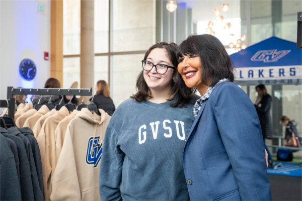 A women in a GVSU sweatshirt, and a woman in a suit jacket pose together looking toward a camera that is to the left out of frame. 