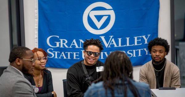 group of four students talk to Koleta Moore, who has her back to the camera, GVSU flag as backdrop