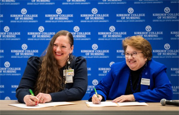 two people smiling and signing papers in front of a KCON backdrop