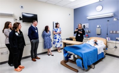 people in simulation hospital room with mannequin, person in black scrubs near bedside talking