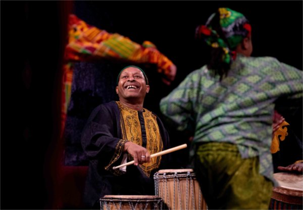 A drummer wearing cultural clothing smiles as young dancers circle.  
