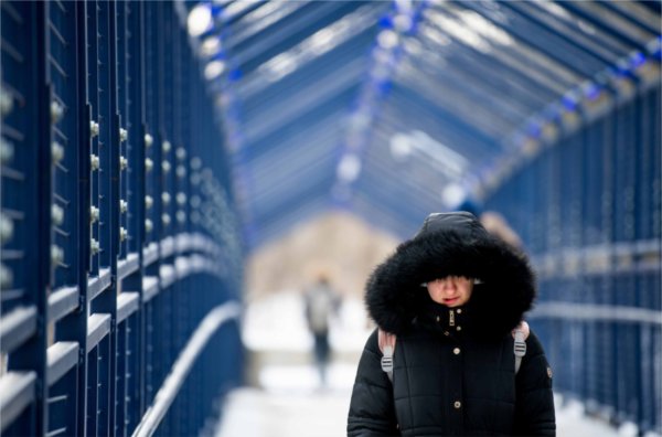  A college student walks across a blue metal bridge while their face is concealed by a large winter coat and hood.