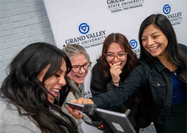  Four people all laugh as they look at a computer screen showing photos of themselves. 