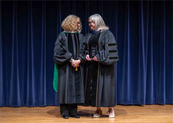  Two faculty members in academic regalia stand together while one holds an award. 