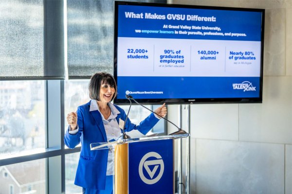A person standing a podium smiles while gesturing with their hands. The screen behind them contains statistics saying 22,000+ students and 90% of graduates employed or in further education.