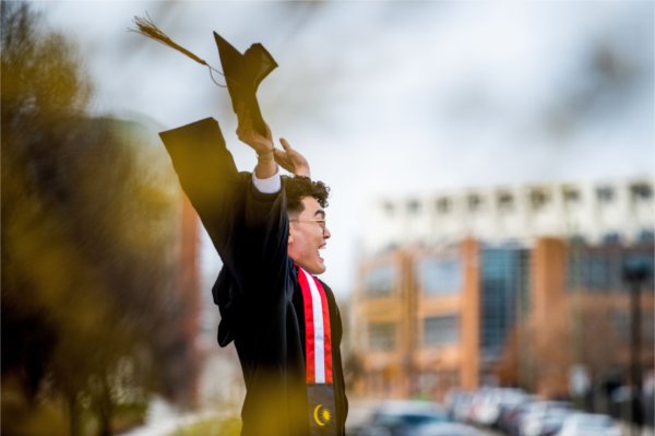 A graduate student throws their arms up in excitement holding a graduation cap and wearing a black graduation gown. 