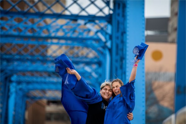  Two college student embrace and toss their arms in the air as they wear blue caps and gowns. 