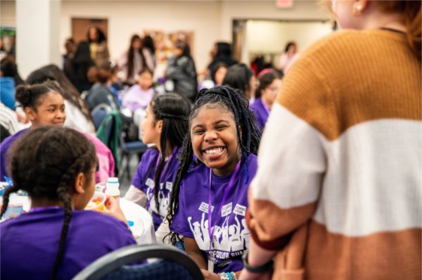 A student wearing a purple shirt smiles among a room full of event participants. 