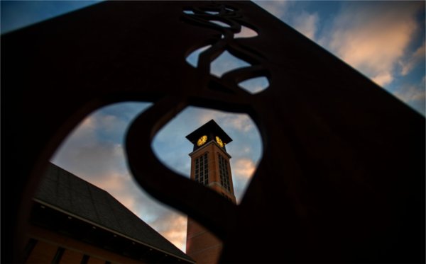 A carillon tower is seen through the shapes of a metal sculpture during sunrise. 